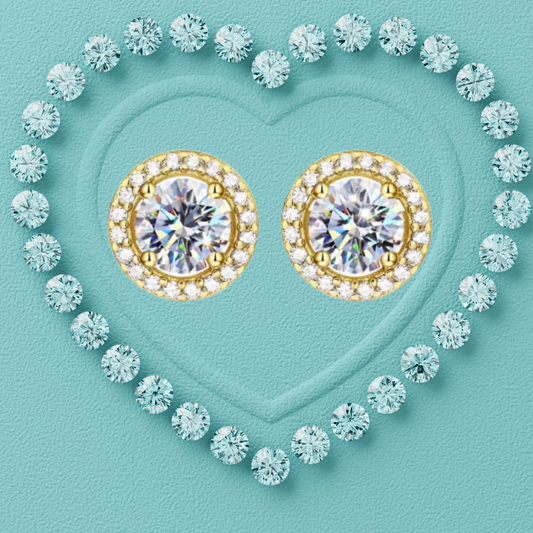 A pair of gold stud earrings with large round clear center stones surrounded by small clear stones, arranged inside a heart outline made of light blue gemstones on a turquoise background.