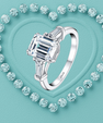 A silver ring with a large emerald-cut clear center stone and baguette side stones, displayed on a turquoise heart-shaped background surrounded by small matching stones.