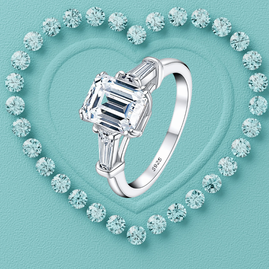 A silver ring with a large emerald-cut clear center stone and baguette side stones, displayed on a turquoise heart-shaped background surrounded by small matching stones.