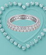 A silver eternity ring paved with clear round-cut gemstones, centered on a textured light blue background with a heart-shaped outline of matching stones.