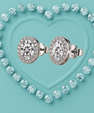 A pair of round diamond halo stud earrings in silver-toned metal, arranged side by side on a textured turquoise background with a heart outline made of small diamonds.