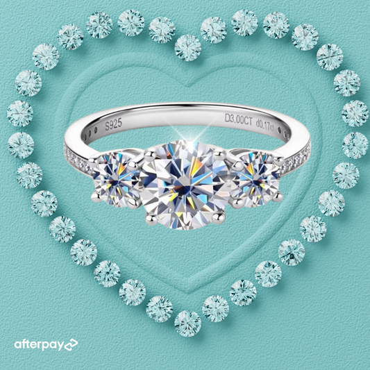 A 'Eternally Yours' 3 Stone Moissanite Ring with a large central moissanite flanked by two smaller ones, set in a heart-shaped arrangement with diamond-like stones on a blue background.