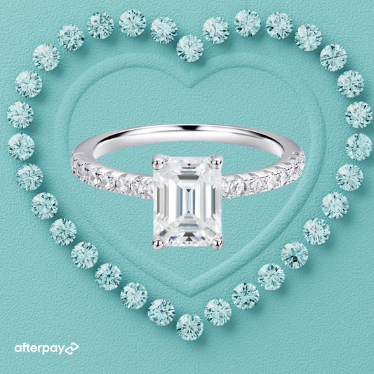 An emerald-cut moissanite solitaire ring with a pavé band, displayed on a heart-shaped background.