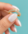 A close-up of a hand with long, glossy white manicured nails holding a gold ring adorned with pave-set clear stones against a solid turquoise background.
