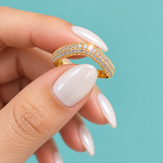 A close-up of a hand with long, glossy white manicured nails holding a gold ring adorned with pave-set clear stones against a solid turquoise background.