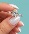A close-up of a hand with iridescent white nail polish holding a silver engagement ring with an oval-cut clear center stone and smaller side stones, set against a solid teal background.