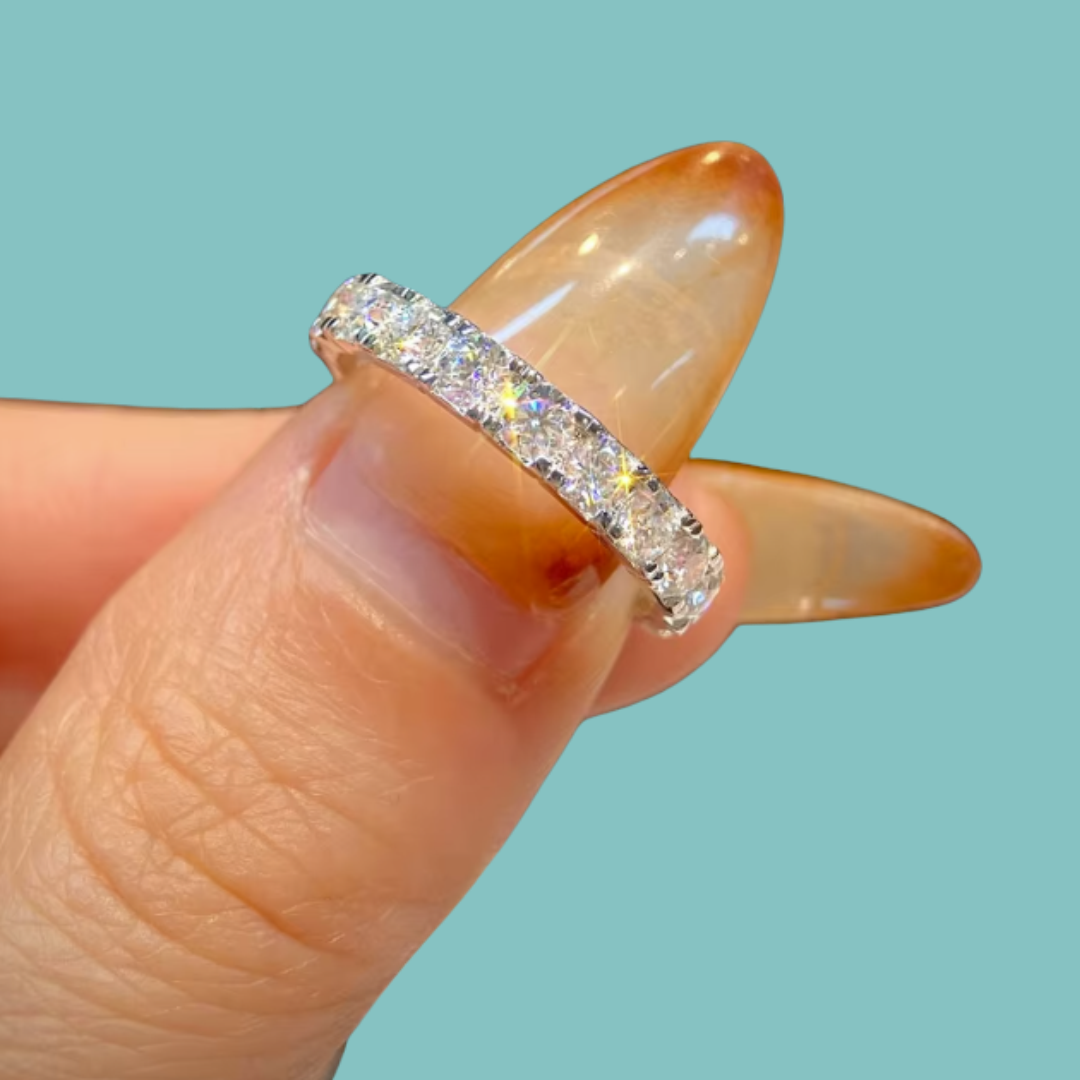 A close-up of a diamond eternity ring on a person's finger, featuring a silver band set with clear round-cut diamonds, set against a solid light teal background.