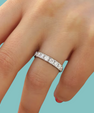 A close-up of a hand wearing a silver diamond eternity ring, set against a solid light teal background.