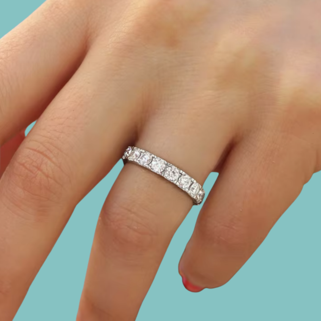 A close-up of a hand wearing a silver diamond eternity ring, set against a solid light teal background.