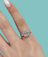 An emerald-cut diamond engagement ring with baguette side stones set in a silver-toned band, shown worn on a hand against a solid teal background.