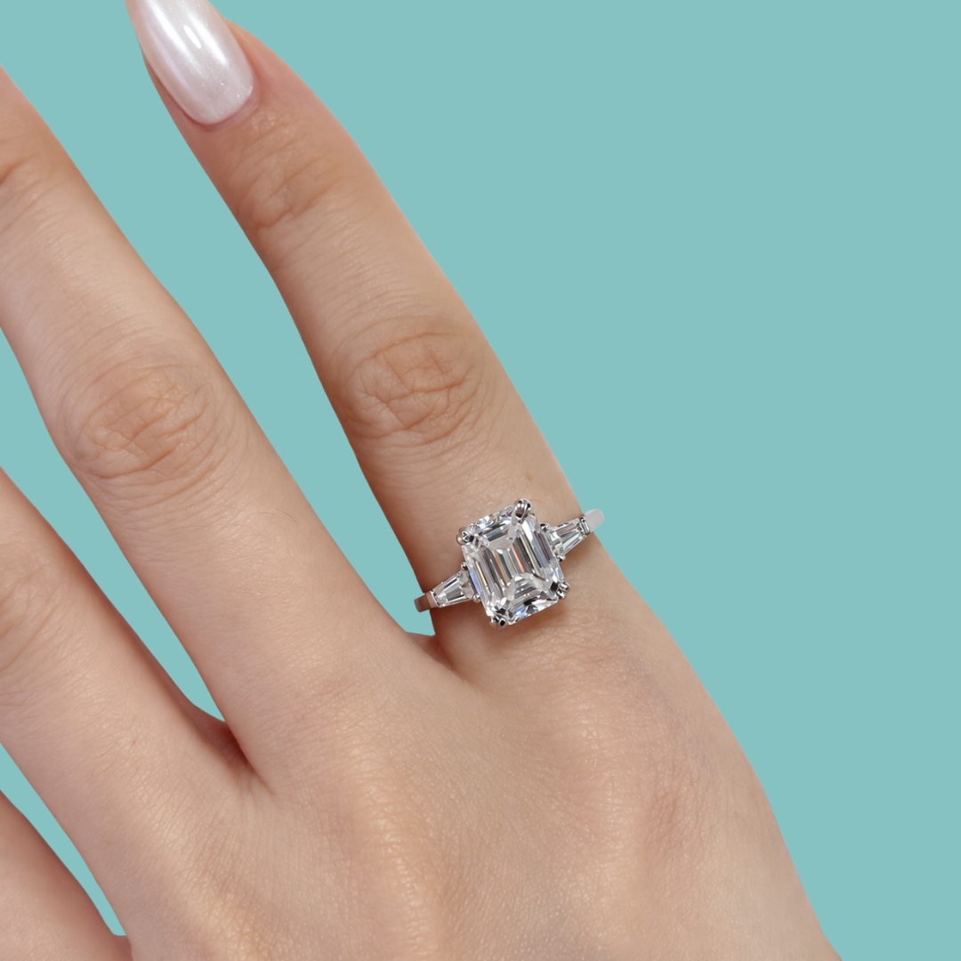 An emerald-cut diamond engagement ring with baguette side stones set in a silver-toned band, shown worn on a hand against a solid teal background.