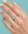 A close-up of a hand with pale pink manicured nails wearing an emerald-cut diamond ring on a light blue background.