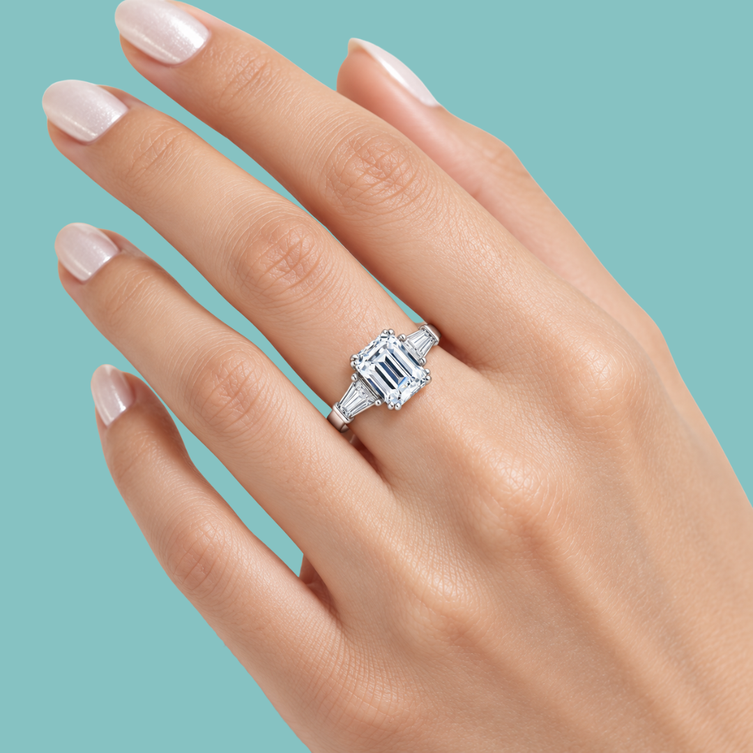 A close-up of a hand with pale pink manicured nails wearing an emerald-cut diamond ring on a light blue background.