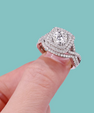 A close-up of a halo diamond ring with a square-cut center stone and surrounding pavé bands, shown being worn on a finger against a solid teal background.