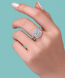 A close-up of a hand wearing a silver-toned halo ring with a round-cut center stone and surrounding pavé stones, set against a solid teal background.