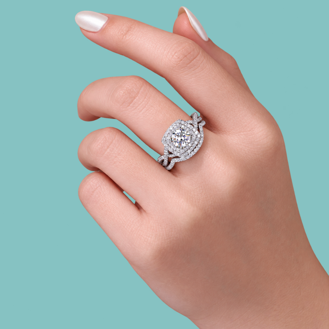A close-up of a hand wearing a silver-toned halo ring with a round-cut center stone and surrounding pavé stones, set against a solid teal background.