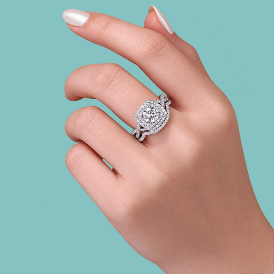 A close-up of a hand wearing a silver-toned halo ring with a round-cut center stone and surrounding pavé stones, set against a solid teal background.