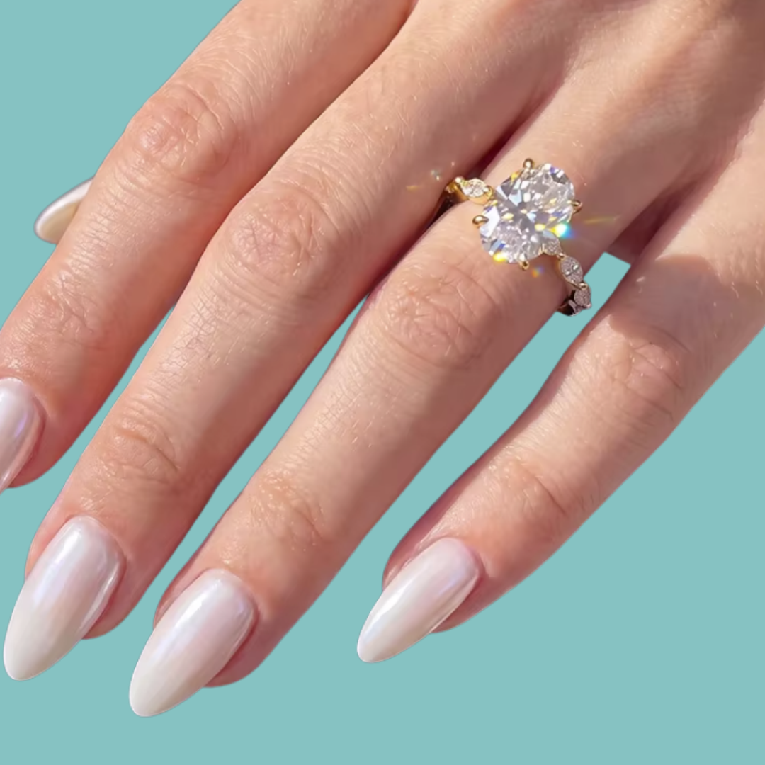 A close-up of a hand with long, glossy pale pink manicured nails wearing a gold solitaire engagement ring with a large clear round gemstone, set against a solid light teal background.