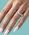 A close-up of a hand with long white manicured nails wearing a silver pavé band ring with sparkling clear stones, set against a solid teal background.