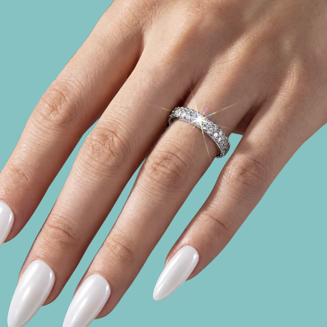 A close-up of a hand with long white manicured nails wearing a silver pavé band ring with sparkling clear stones, set against a solid teal background.