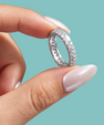 A close-up of a hand with long, manicured nails holding a silver eternity ring paved with small clear stones against a solid teal background.