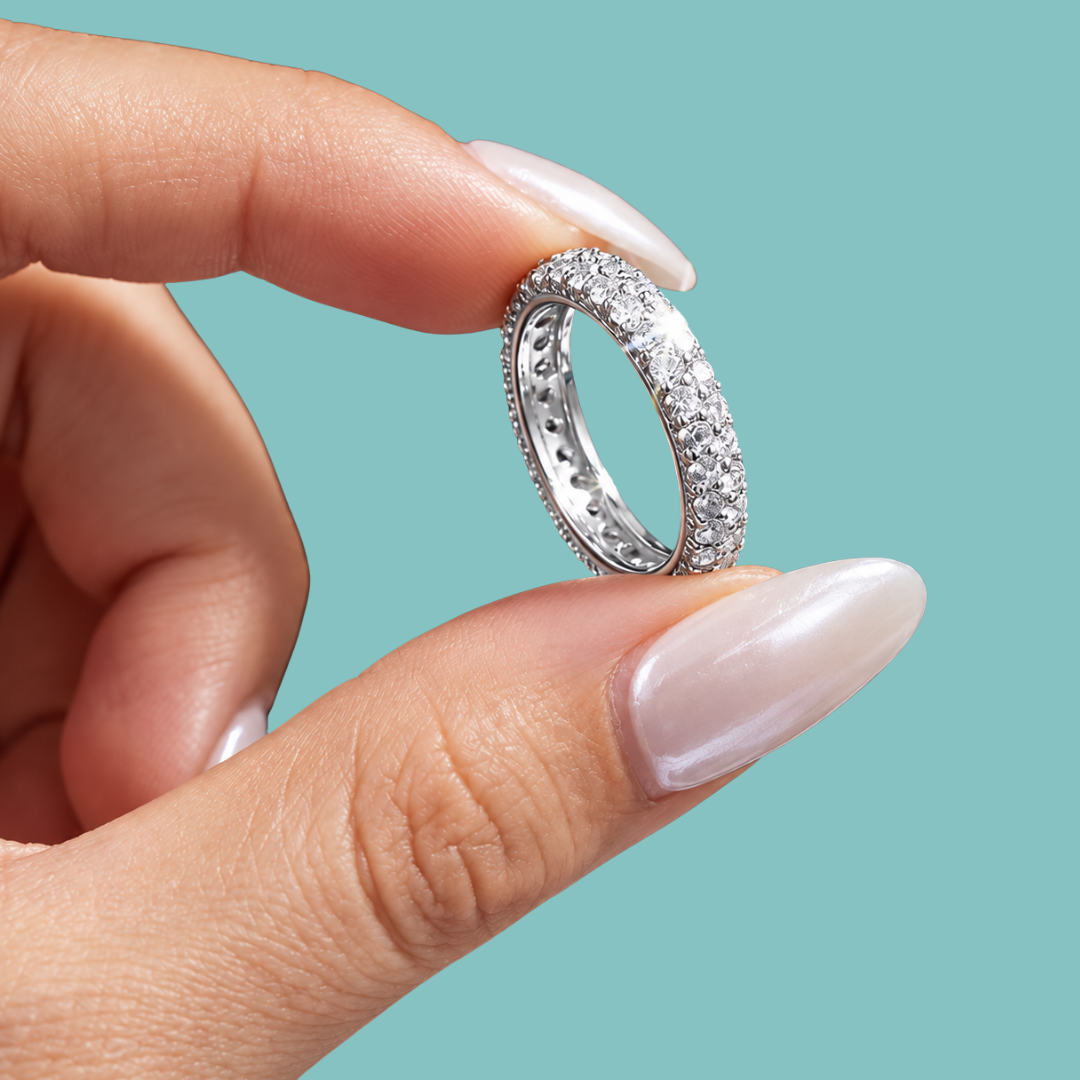 A close-up of a hand with long, manicured nails holding a silver eternity ring paved with small clear stones against a solid teal background.