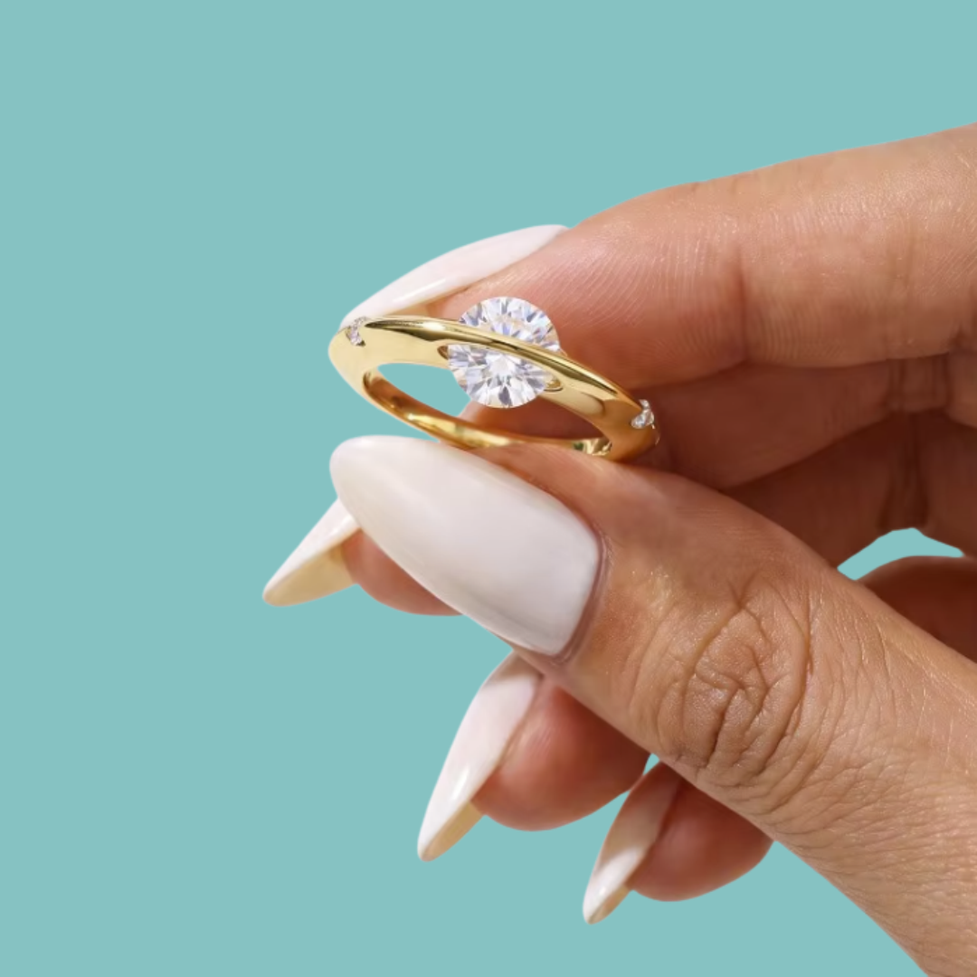 Saturn’s Kiss moissanite ring featuring a round centre stone crossed by a polished gold-toned band, with small side stones.