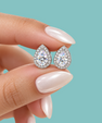 A close-up of a hand with pale pink manicured nails holding a pair of teardrop-shaped halo stud earrings with clear round-cut center stones and small surrounding stones, set against a solid light aqua background.