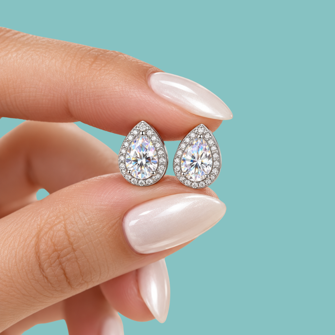A close-up of a hand with pale pink manicured nails holding a pair of teardrop-shaped halo stud earrings with clear round-cut center stones and small surrounding stones, set against a solid light aqua background.