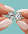 A close-up of a person's hands with long, pale pink manicured nails holding a pair of oval halo stud earrings. The earrings feature round brilliant-cut center stones surrounded by a border of smaller stones in a silver-toned setting, set against a solid light teal background.