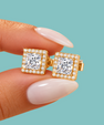 A close-up of a hand with long, pale pink manicured nails holding a pair of gold square stud earrings with princess-cut clear gemstones and halo accents, set against a solid teal background.