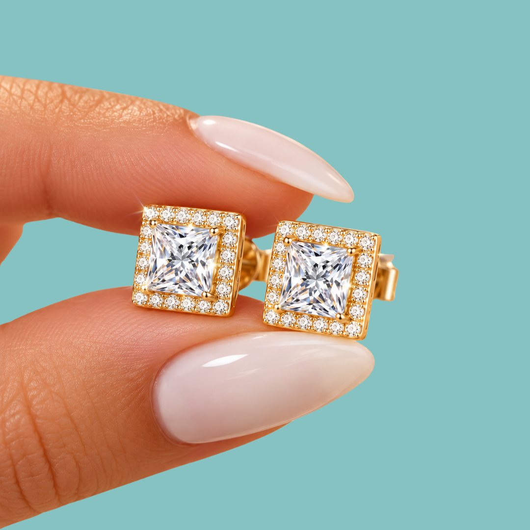 A close-up of a hand with long, pale pink manicured nails holding a pair of gold square stud earrings with princess-cut clear gemstones and halo accents, set against a solid teal background.