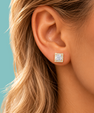 A close-up of a woman's ear wearing a square stud earring with a clear central gemstone surrounded by a halo of small gold and white stones, set against a solid teal background.