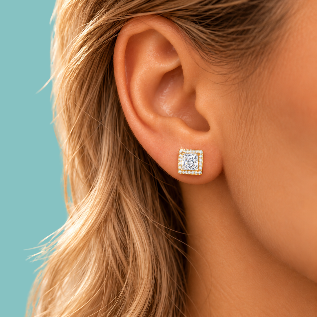 A close-up of a woman's ear wearing a square stud earring with a clear central gemstone surrounded by a halo of small gold and white stones, set against a solid teal background.