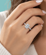 A close-up of a hand wearing a silver engagement ring with a double-stone emerald-cut center and a pavé band, set against a soft white knit sweater and a teal background.