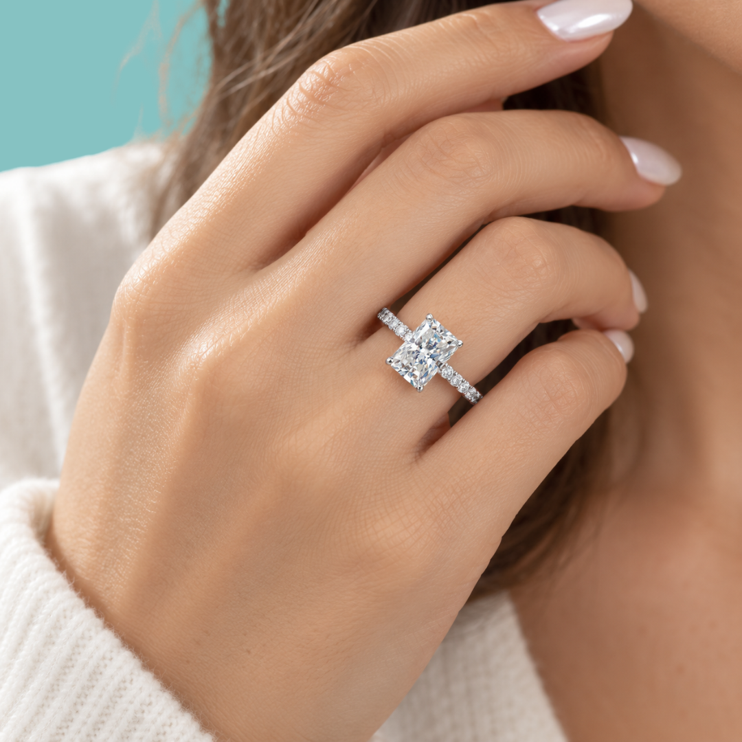A close-up of a hand wearing a silver engagement ring with a double-stone emerald-cut center and a pavé band, set against a soft white knit sweater and a teal background.
