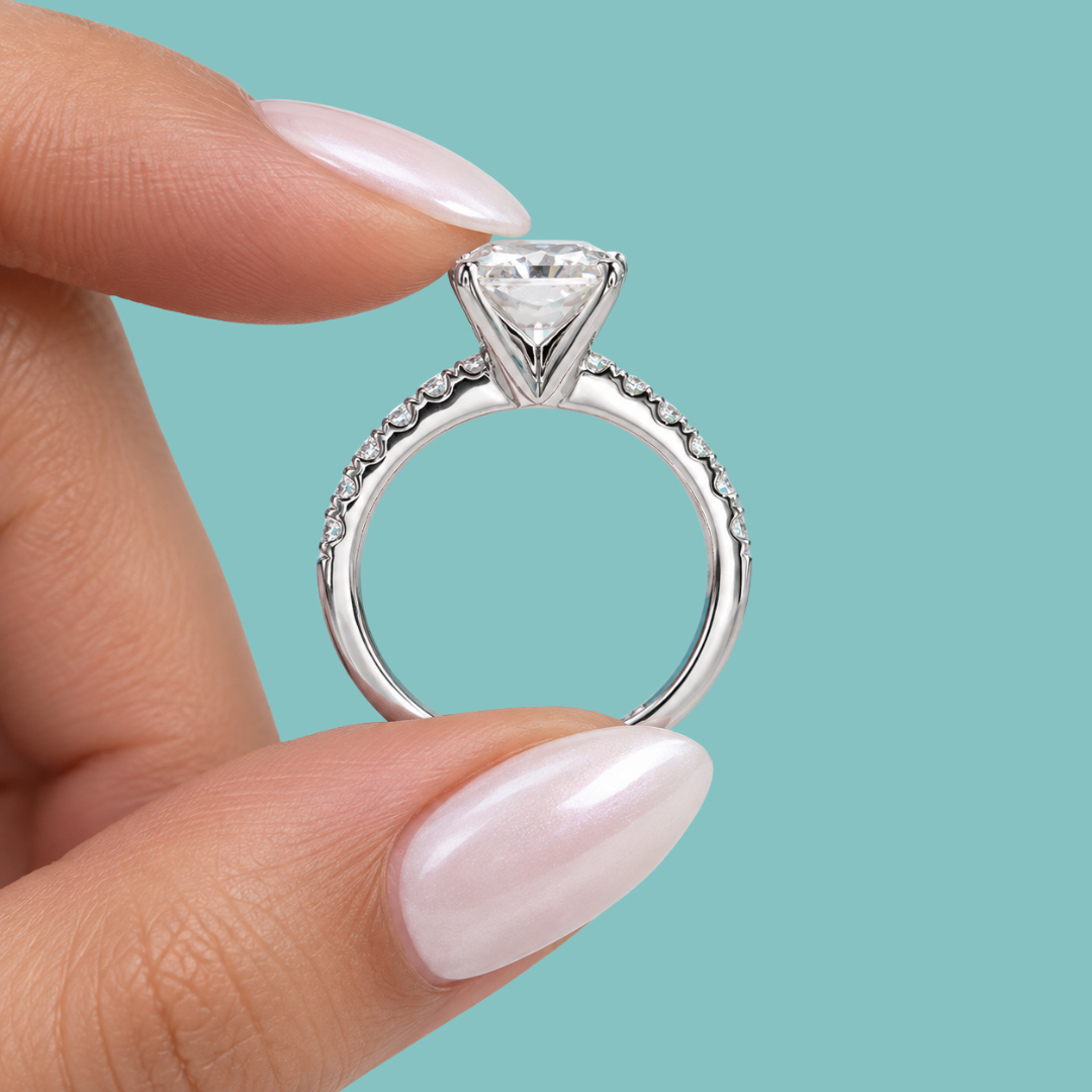 A close-up of a hand with pale pink manicured nails holding a silver engagement ring with a triangular clear gemstone and small accent stones on the band, set against a solid teal background.