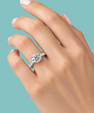 A close-up of a hand wearing a silver-toned diamond engagement ring with a round-cut center stone and pavé band, set against a solid teal background.