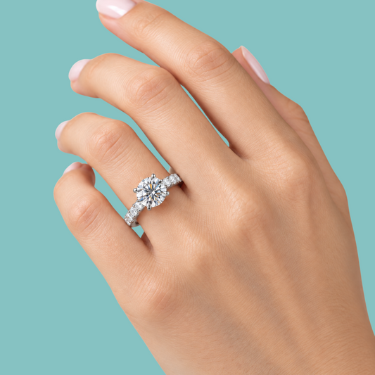 A close-up of a hand wearing a silver-toned diamond engagement ring with a round-cut center stone and pavé band, set against a solid teal background.