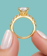 A close-up of a gold solitaire engagement ring with a cushion-cut diamond and a pavé band, held between two fingers against a solid teal background.
