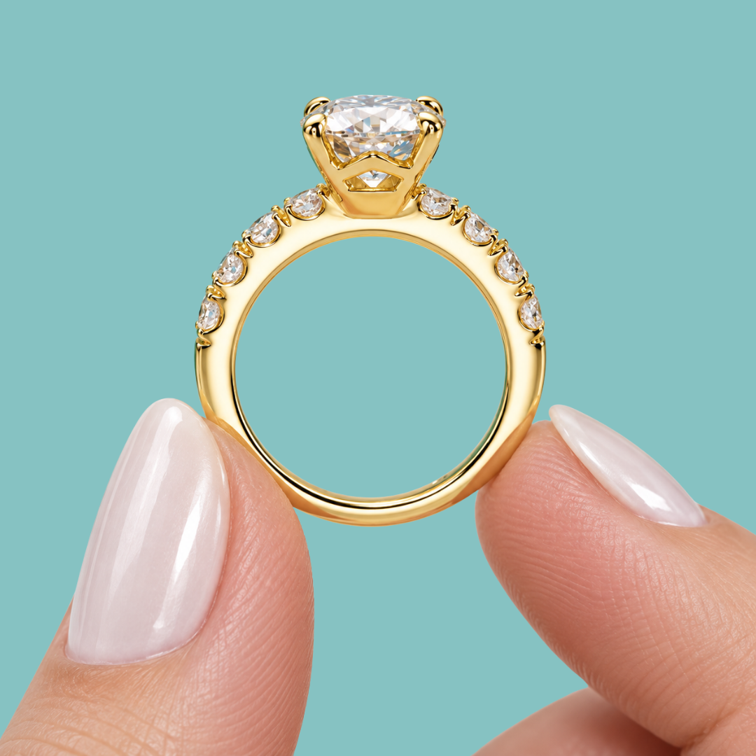 A close-up of a gold solitaire engagement ring with a cushion-cut diamond and a pavé band, held between two fingers against a solid teal background.