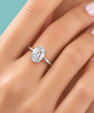 A close-up of a hand wearing a silver solitaire ring with an oval-cut clear gemstone, set against a solid teal background.