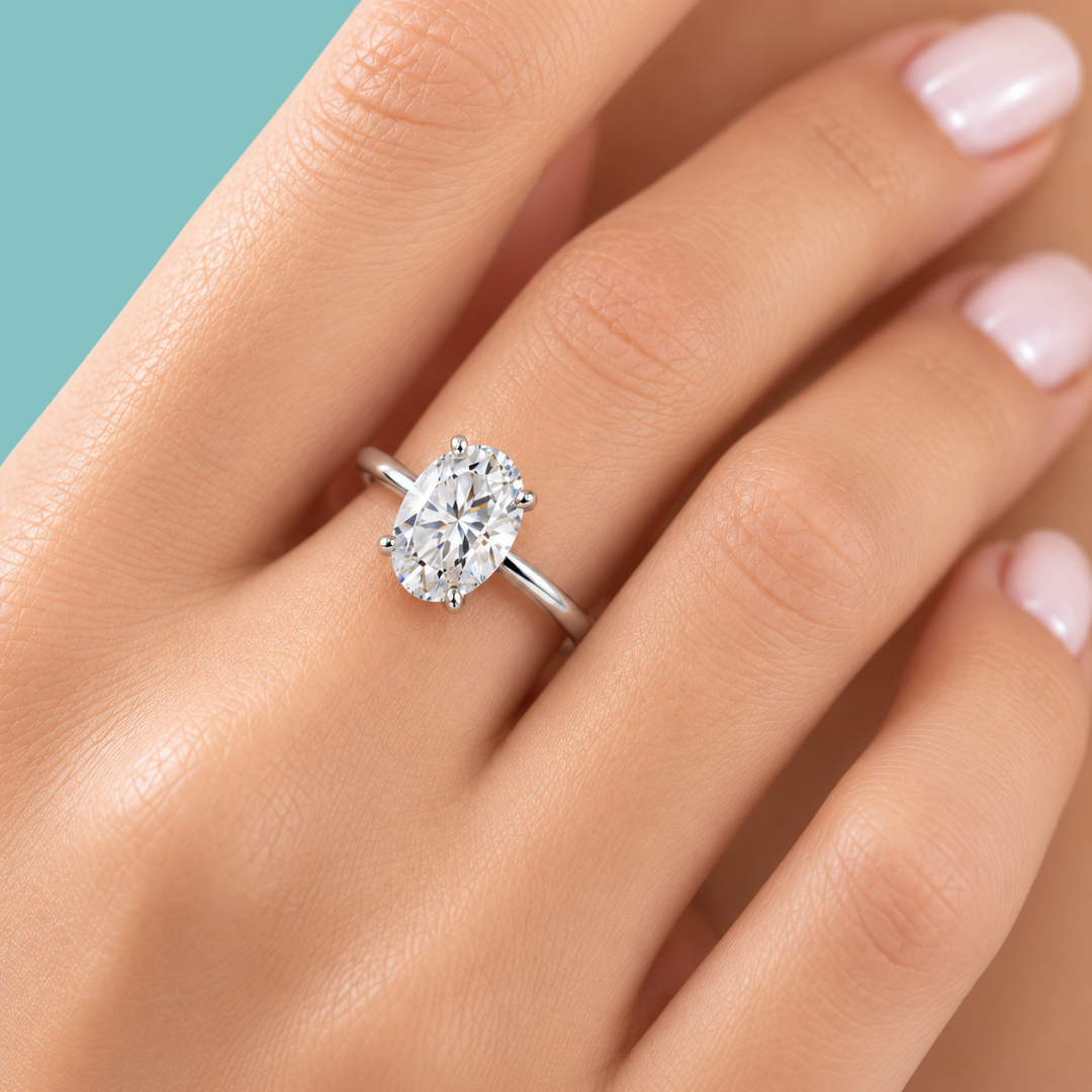 A close-up of a hand wearing a silver solitaire ring with an oval-cut clear gemstone, set against a solid teal background.
