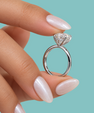 A close-up of a hand with long, glossy pale pink manicured nails holding a silver-toned diamond ring with a heart-shaped clear gemstone against a solid light blue background.