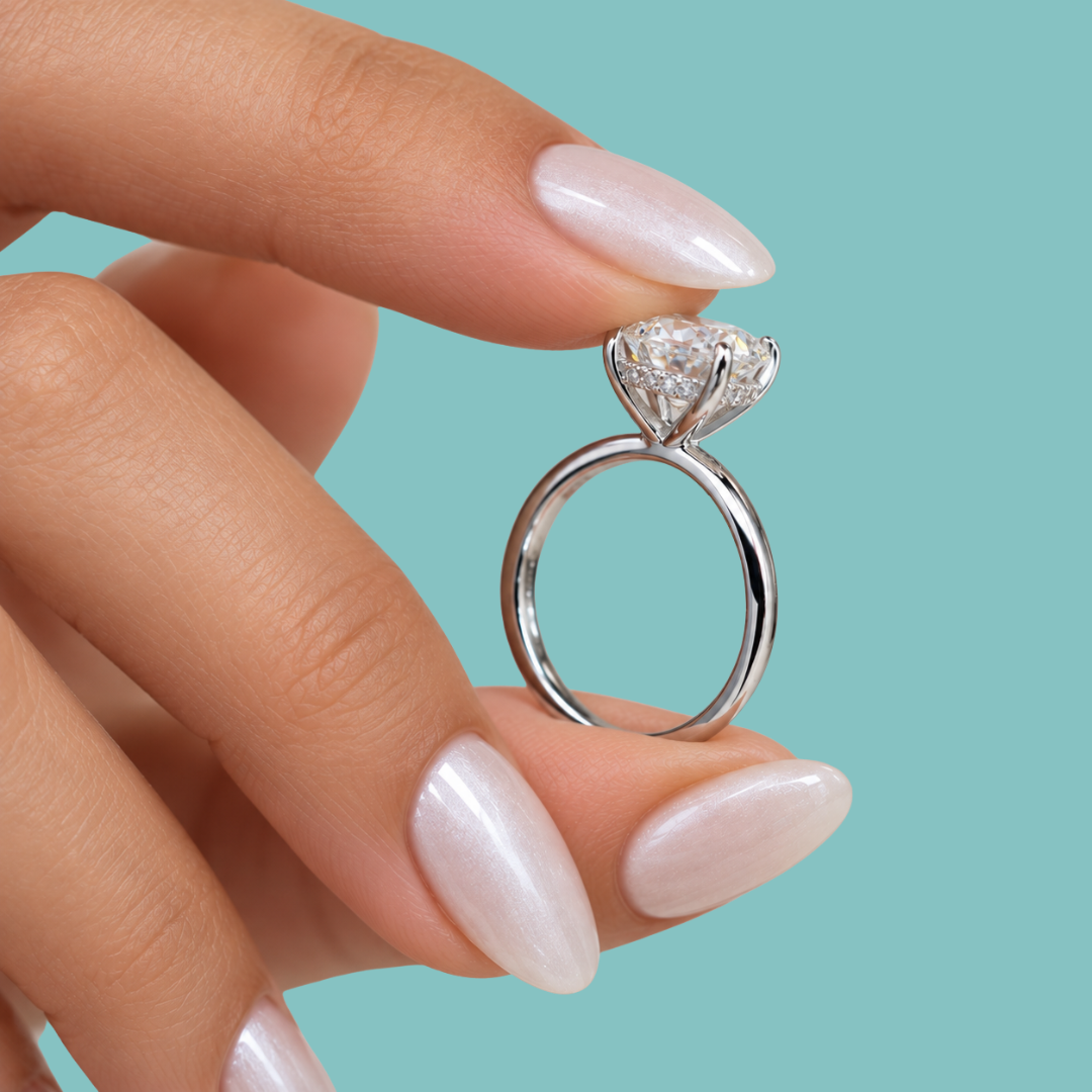 A close-up of a hand with long, glossy pale pink manicured nails holding a silver-toned diamond ring with a heart-shaped clear gemstone against a solid light blue background.