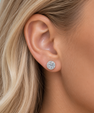 A close-up of a woman's ear wearing a round stud earring with a halo of small clear stones in a silver-toned setting.