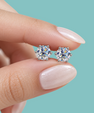 A close-up of a person's hand with pale pink manicured nails holding a pair of round-cut solitaire stud earrings with silver settings against a solid light teal background.