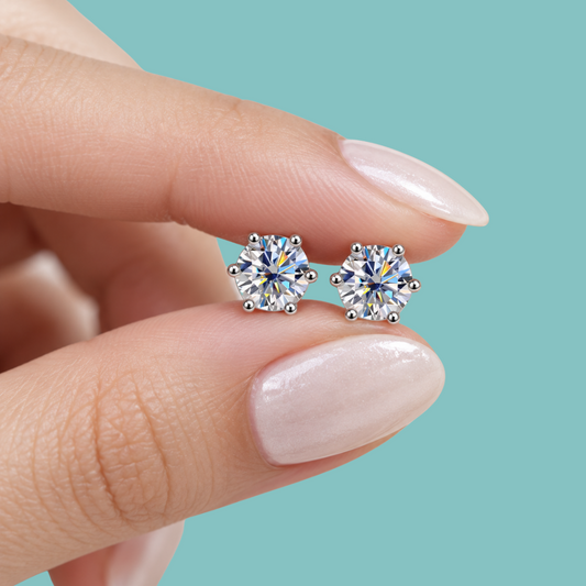 A close-up of a person's hand with pale pink manicured nails holding a pair of round-cut solitaire stud earrings with silver settings against a solid light teal background.