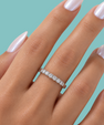 A close-up of a hand with long, glossy pale pink manicured nails wearing a silver eternity ring set with clear stones, set against a solid teal background.