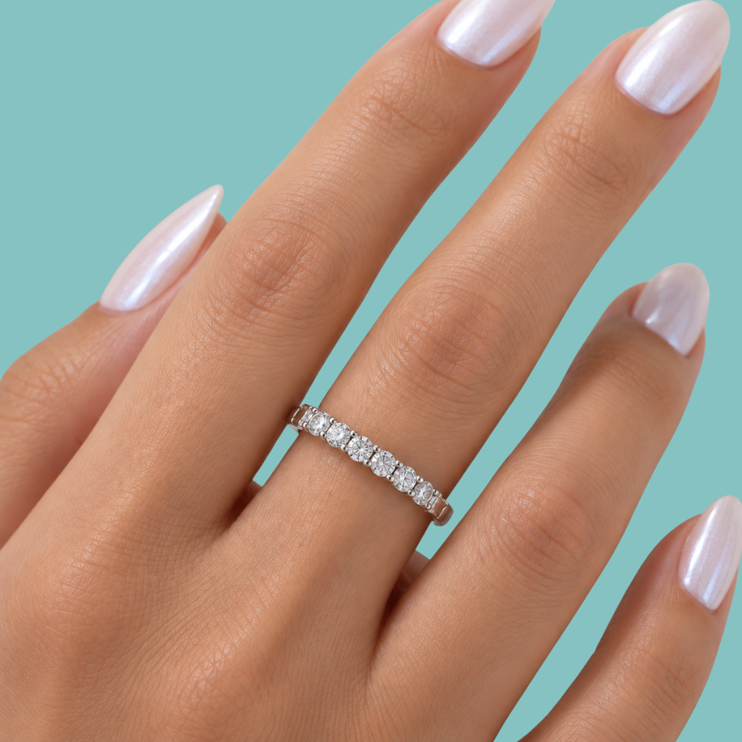 A close-up of a hand with long, glossy pale pink manicured nails wearing a silver eternity ring set with clear stones, set against a solid teal background.